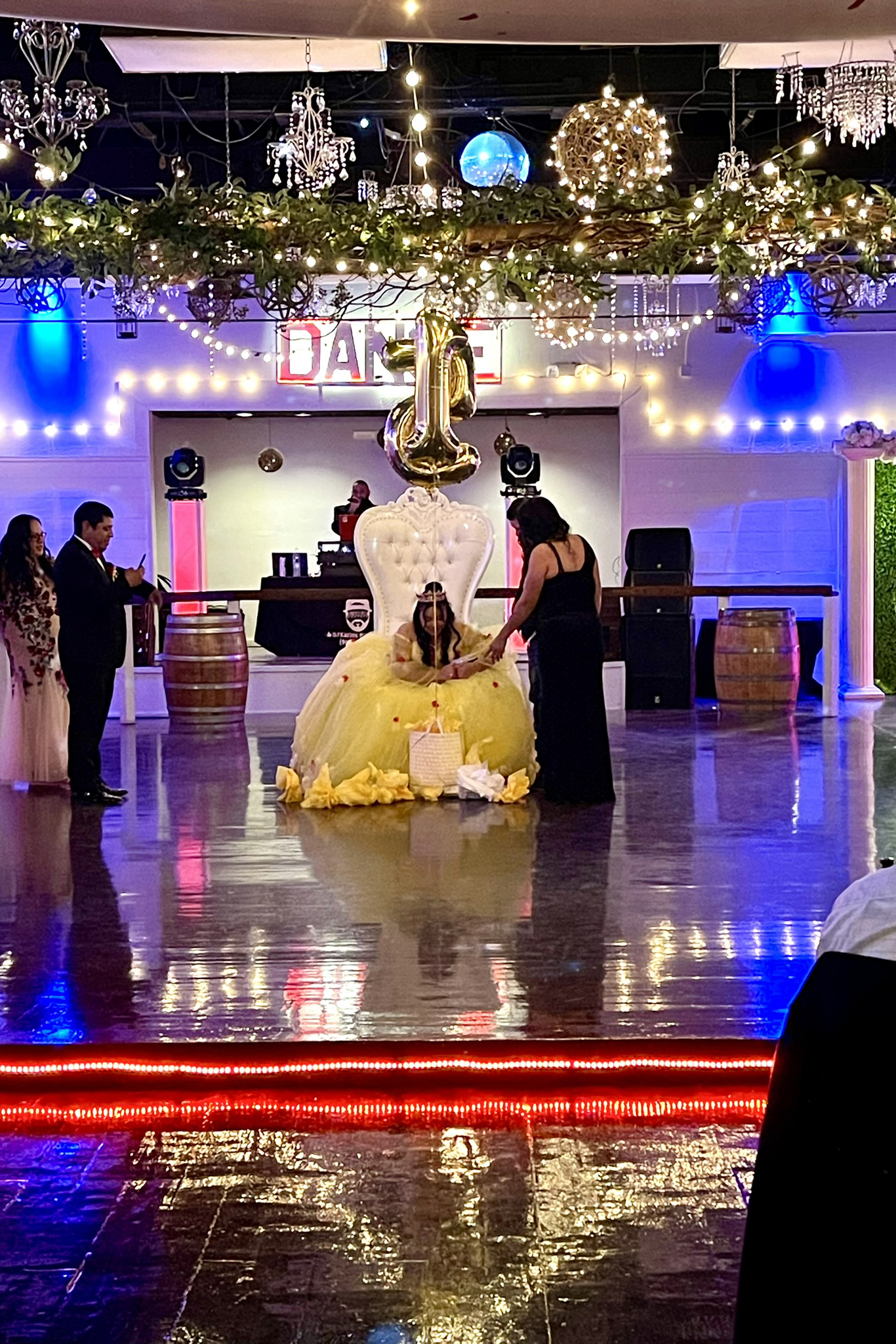Quinceanera girl on illuminated dance floor in chair of honor receiving gifts.